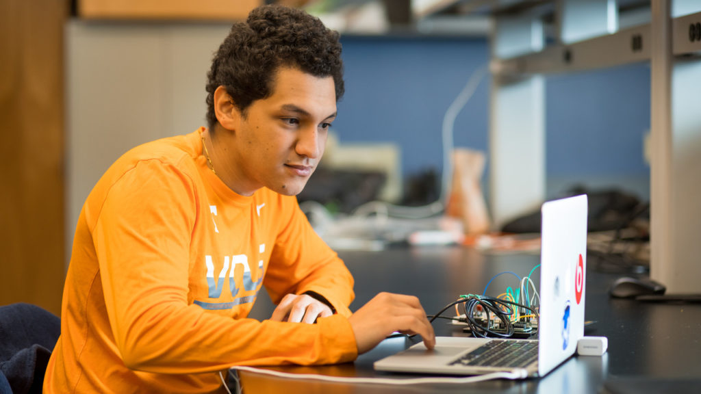 A student wearing an orange Tennessee shirt sitting down using his laptop.