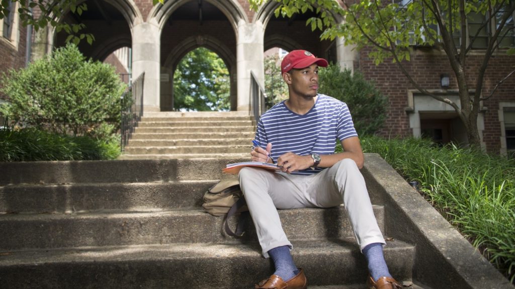 A student wearing a blue shirt and red hat sitting outside on some stairs writing in a notebook on campus.