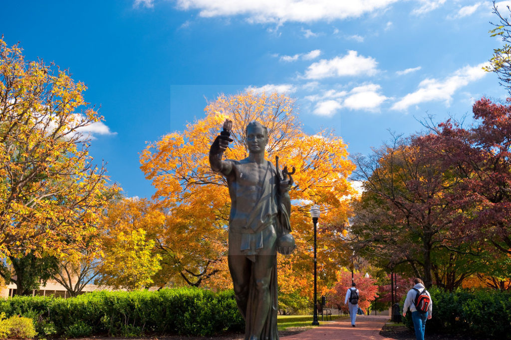 The torch bearer on campus showing fall colors on the trees in Circle Park. There are two students walking with their backpacks on in the background as well.